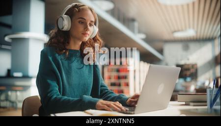 Concentrated Young Female Student With Headphones Using Laptop In Modern Library. Caucasian Woman Engaged In Online Learning, Surrounded By Books, Focused On Her Academic Research. Stock Photo