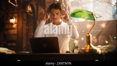Serious Young Man Studying With Laptop in a Classical Library, Wearing Headphones and Focused on E-Learning. Caucasian Male Engaged in Academic Research Surrounded by Classical Architecture and Books Stock Photo