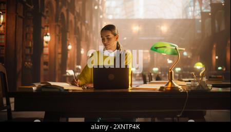 Dedicated Female Student Engaged in E-Learning at a Classic Library Desk. Young Woman Using Laptop for Research, Surrounded by Books, in a Classical Library Setting with Ornate Architecture. Stock Photo
