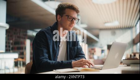 Concentrated Young Caucasian Male Student Engaged in Academic Research With Laptop and Books in Modern Library. Man Studying Hard, Focused on Computer Screen, Surrounded by Shelves Full of Books. Stock Photo