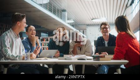 Diverse Group of Students Collaborating on a Project in a Modern Library. Young Adults Engaged in Discussion, Using Laptops and Books, Sharing Ideas and Laughing Together. Stock Photo