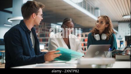 Diverse Group of Students Collaborating on a Project in a Modern Library. Young Black Female and Caucasian Peers Discussing Academic Materials and Using a Laptop for Research. Stock Photo