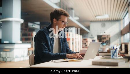 Young Man Studying in Modern Library. Caucasian Male Using Laptop Computer for College Research, Surrounded by Books and Academic Materials, Focused on Learning and Education in University Setting Stock Photo