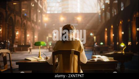 Serene Female Student Engaged in Online Learning in a Classical Library. Young Woman Wearing Headphones, Using Laptop and Books to Study Amidst Classical Architecture and Wooden Furniture. Stock Photo