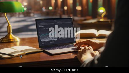 Analytical Business Professional Reviewing Financial Data on a Laptop in a Classic Library. Person Analyzing Market Trends and Graphs, Surrounded by Books and a Green Desk Lamp. Stock Photo