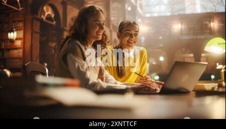 Joyful Caucasian and Hispanic Female Students Collaborating on a Project in a Vintage Library. They are Using a Laptop and Taking Notes, Surrounded by Books and Educational Atmosphere Stock Photo