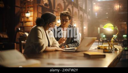 Engaged Multiethnic Students Discussing Academic Project in Classical Library. Young Black Female and Caucasian Male Using Laptop for Research, Surrounded by Books and Vintage Decor. Stock Photo