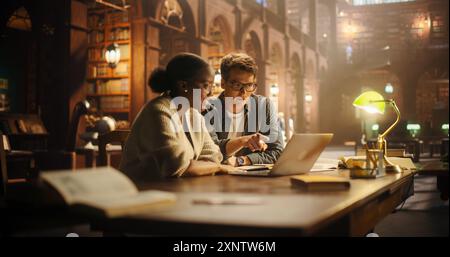 Engaged Multiethnic Students Collaborating on a Project in a Historic Library. Young Man and Woman Using Laptop, Discussing Academic Research Surrounded by Books in a Warmly Lit Study Environment. Stock Photo