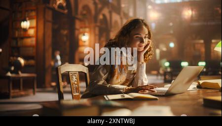 Radiant Young Woman Engaged in Online Education, Using Laptop in Classical Library. Curly-Haired Student Researches for College Project Amidst Antique Books, Illuminated by Warm, Ambient Light Stock Photo