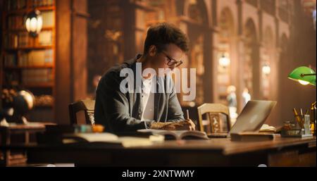 Concentrated Young Caucasian Male Student Engaged in Academic Research in a Classic Library, Using Laptop and Books for Studying. Scholar Writing Notes, Surrounded by Wooden Shelves Filled with Books. Stock Photo