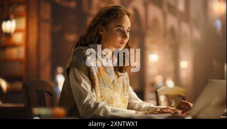 Serene Young Woman Studying With Laptop in a Vintage Library, Surrounded by Books and Warm Lighting. Focused on E-Learning, She Engages in Academic Research and Note-Taking in a Cozy Setting. Stock Photo