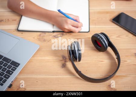 Detail shot of a desk where a hand is writing on a notepad. Above the desk are headphones, a smartphone and a laptop. Stock Photo
