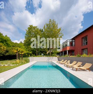 Red-walled villa in a big green garden with trees. There is a big swimming pool and sun loungers. Frontal view. Sunny summer day. Stock Photo