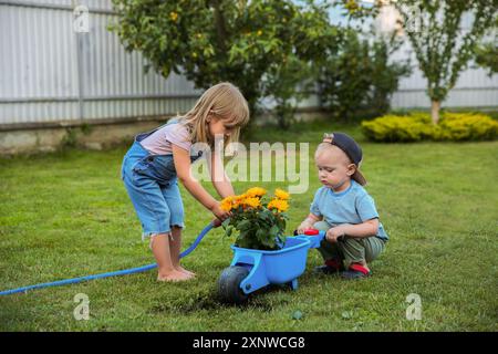 Cute little boy holding wheelbarrow while his sister watering flowers outdoors Stock Photo