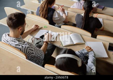 High angle shot of disinterested students sitting at desk with notebooks and mobile phones distracting from lecture at university class Stock Photo