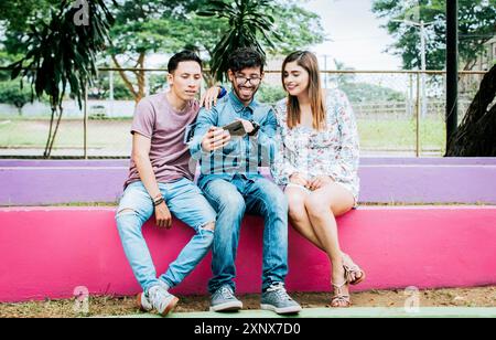 Three happy friends looking at the cell phone sitting in the park. Three happy people looking at cell phone outdoors Stock Photo