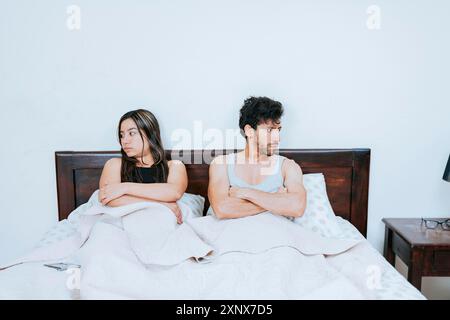 Conflicting couple sitting on bed, Upset couple sitting on bed with crossed arms. Young couple arguing sitting in bed, Upset couple sitting on the bed Stock Photo