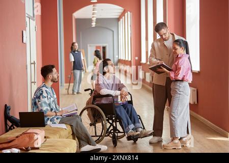 Diverse group of students asking adult male professor question during informal conversation in pink university hallway Stock Photo