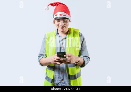 Smiling engineer in christmas hat using cell phone isolated. Cheerful young engineer in christmas hat using smartphone Stock Photo