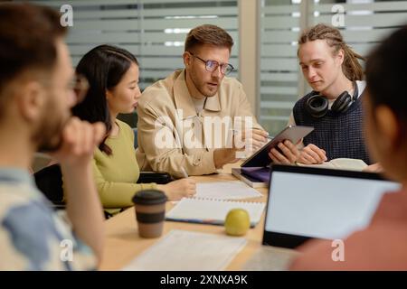 Adult male teacher explaining task to multiethnic college students pointing at digital tablet screen while sitting at desk during group seminar in classroom Stock Photo