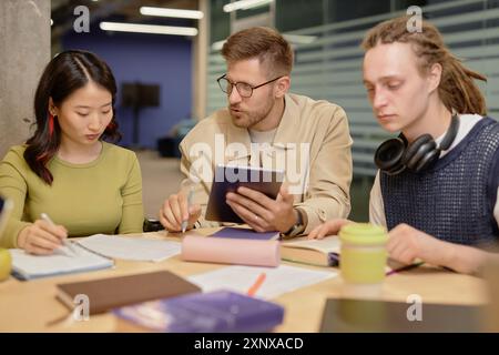 Confident university male professor in glasses explaining written assignment using tablet to young multiethnic students sitting around him at group tutorial Stock Photo