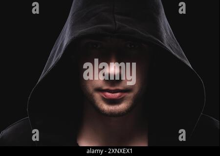 Close-up portrait of a courageous man in a deep dark hood on a black background. The concept of secrecy of secrets and people hiding from the Stock Photo