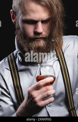 An attractive man with a long bang beard and a mustache sitting against the wall sniffs the scent of an alcoholic beverage in a glass that holds his Stock Photo