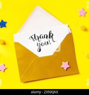 Thank you card in a golden envelope, overhead square shot on a yellow background with glitter stars Stock Photo