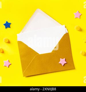 A white greeting card in a golden envelope, overhead square shot on a yellow background with glitter stars Stock Photo