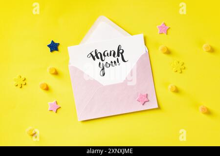 Hand written Thank You card in a pink envelope, overhead shot on a yellow background with glitter stars and candies Stock Photo