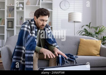 Person wrapped in blanket sitting on couch using heater, coping with cold weather. Man trying to warm up indoors, seeking comfort and warmth at home. Stock Photo