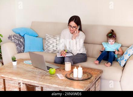 Positive young woman in casual clothes discussing business issue and taking notes in planner while working on remote project in cozy living room near Stock Photo