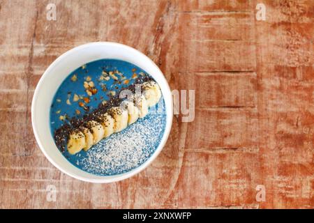 Top view of healthy breakfast bowl with blue smoothie garnished with fresh sliced banana and seeds with coconut flakes on rustic wooden surface Stock Photo