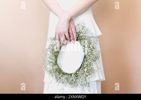 Crop anonymous young female in white wedding dress holding delicate wreath made with white flowers while standing against beige background Stock Photo