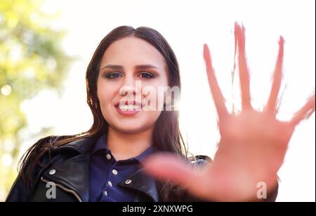 Content young female in leather jacket showing palm at camera and smiling against bright sunlight in park Stock Photo