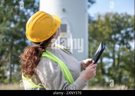 Female engineer with brown curly hair and yellow helmet is looking up a wind turbine while holding a digital tablet Stock Photo