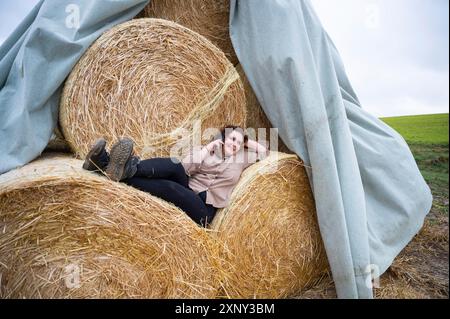 Young woman with brown curly hair is smiling while using her smartphone, lying on a stack of hay balls, country side, side view Stock Photo