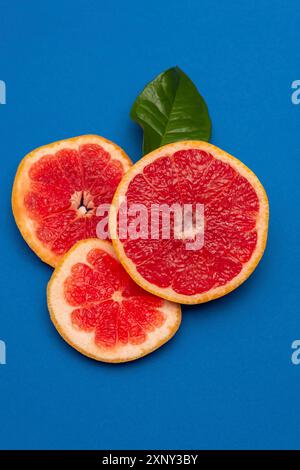 Sliced sides of a grapefruit, three pieces with a green leaf, red pulp isolated on a blue background, vertical shot Stock Photo