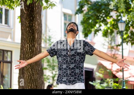 Indian young man taking a deep breath of fresh air, relaxing, taking a break, resting, meditating, calm down outdoors. Hispanic guy enjoying happy day stands on urban city street park. Town lifestyles Stock Photo