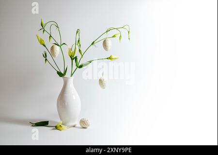 Delicate flower bunch in a white vase decorated with hanging Easter eggs on a light gray background, copy space, selected focus, narrow depth of field Stock Photo