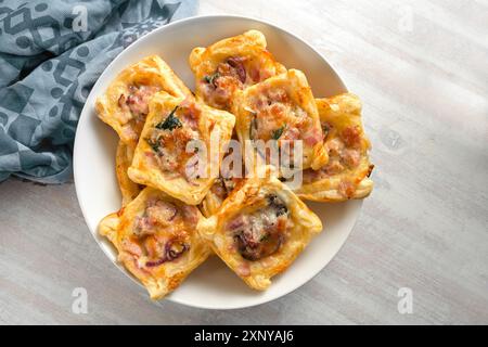 Baked puff pastry squares with ham, onion, herbs and cheese in a bowl, finger food snack for a warm or cold party buffet, high angle view from above Stock Photo