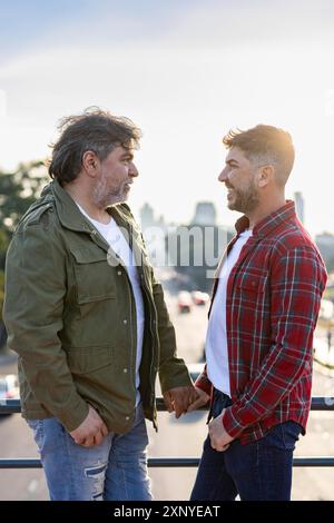 Two men, a gay couple, are standing on a city bridge, holding hands and looking at each other Stock Photo