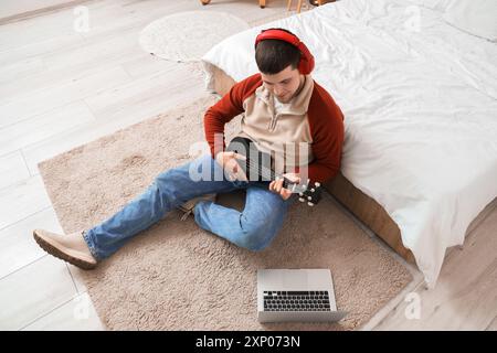 Young bearded man with headphones playing ukulele in bedroom Stock Photo