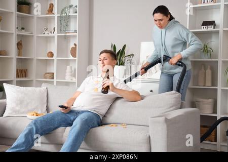 Stressed wife with vacuum cleaner and her lazy husband watching TV at home Stock Photo