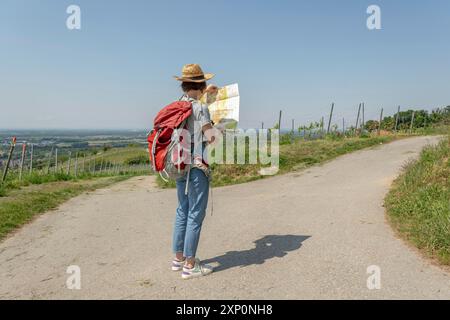 Hiking Woman with brown hair, gray t-shirt, jeans, straw hat and red backpack standing at a country crossroads, looking at a hiking map Stock Photo