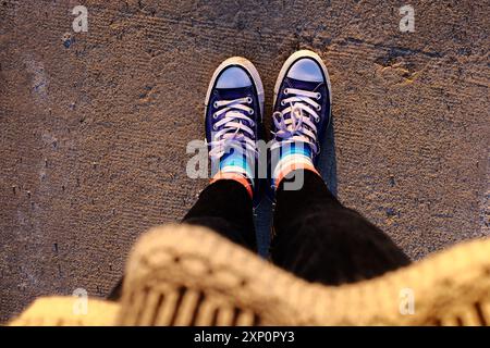 classic blue sneakers with worn white laces, resting concrete ground, shoes with colorful socks. Woman standing to waiting or thinking moment of caref Stock Photo