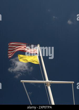 American flag waving over a bright yellow beach warning flag on a white pole with blue skies and a white cloud. Stock Photo