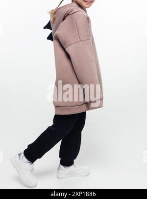 A child walking while wearing a beige hoodie with a black dinosaur tail detail and black pants, smiling against a white background. Stock Photo