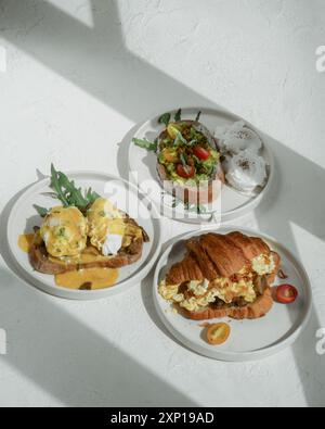 Three plates of brunch dishes: eggs benedict, avocado toast, and a croissant sandwich Stock Photo