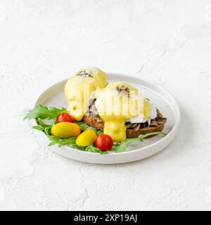Two poached eggs with hollandaise sauce on toasted bread, served with mushrooms and cherry tomatoes Stock Photo